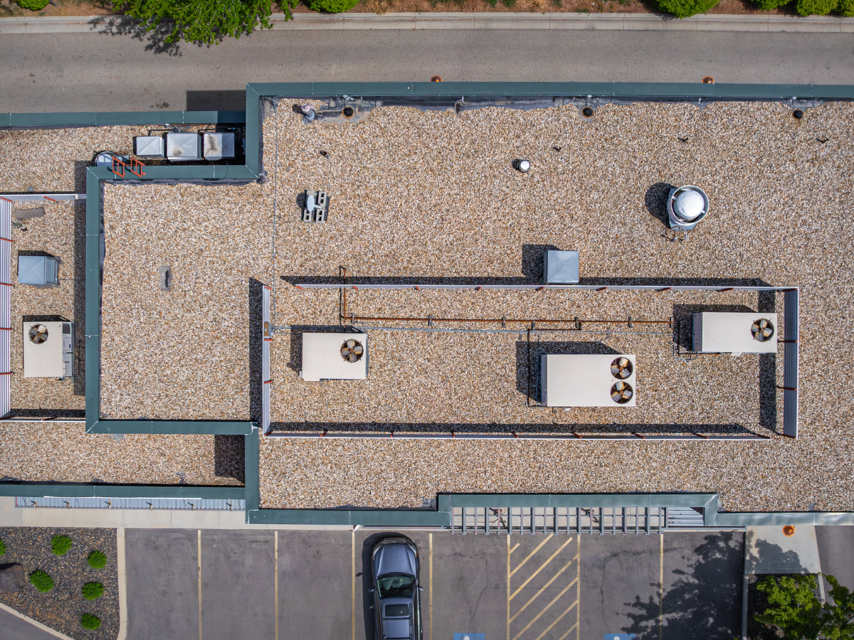 Aerial view of a flat commercial roof covered in gravel, featuring multiple HVAC units and ventilation systems. The rooftop shows signs of aging and wear, with discolored areas and some surface irregularities. A parking lot with one visible vehicle is located at the bottom edge of the image.