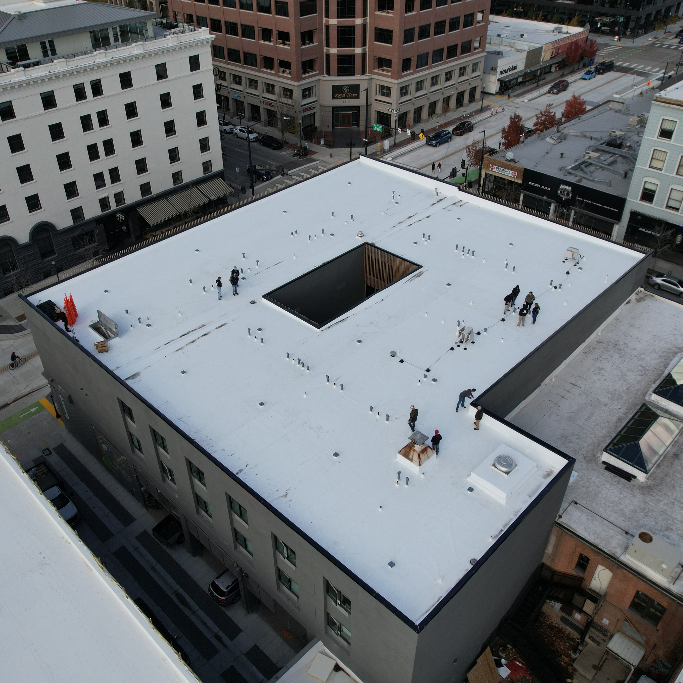 Aerial perspective of a large flat roof on a multi-story commercial building in a downtown area, with workers inspecting the white membrane roofing and a central open courtyard visible.