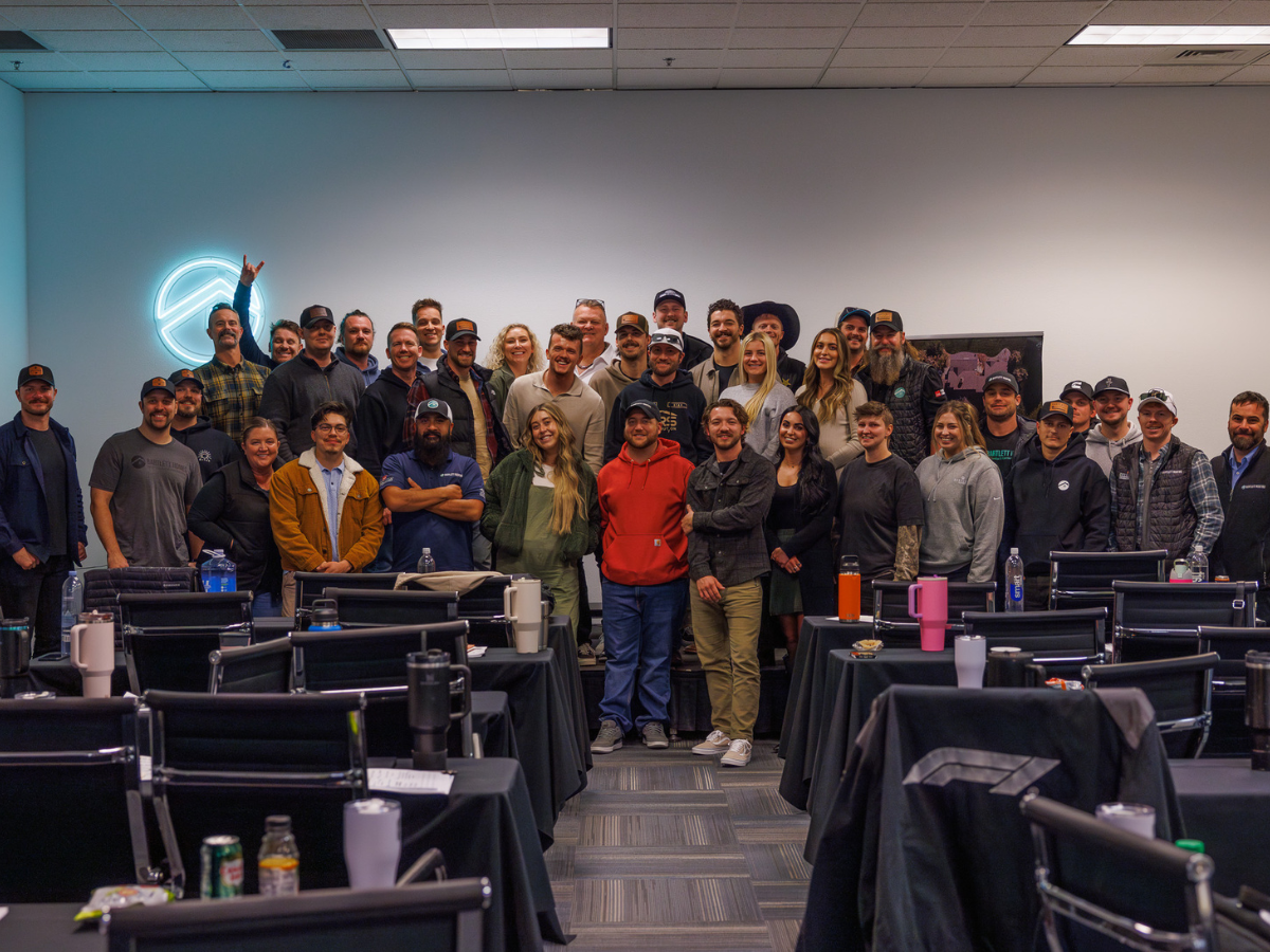 Group photo of over 40 people at a corporate training event, standing together in a conference room with black chairs and tables in the foreground. A neon company logo is lit on the wall to the left, and attendees are smiling and casually dressed, suggesting a team-building or leadership development session.