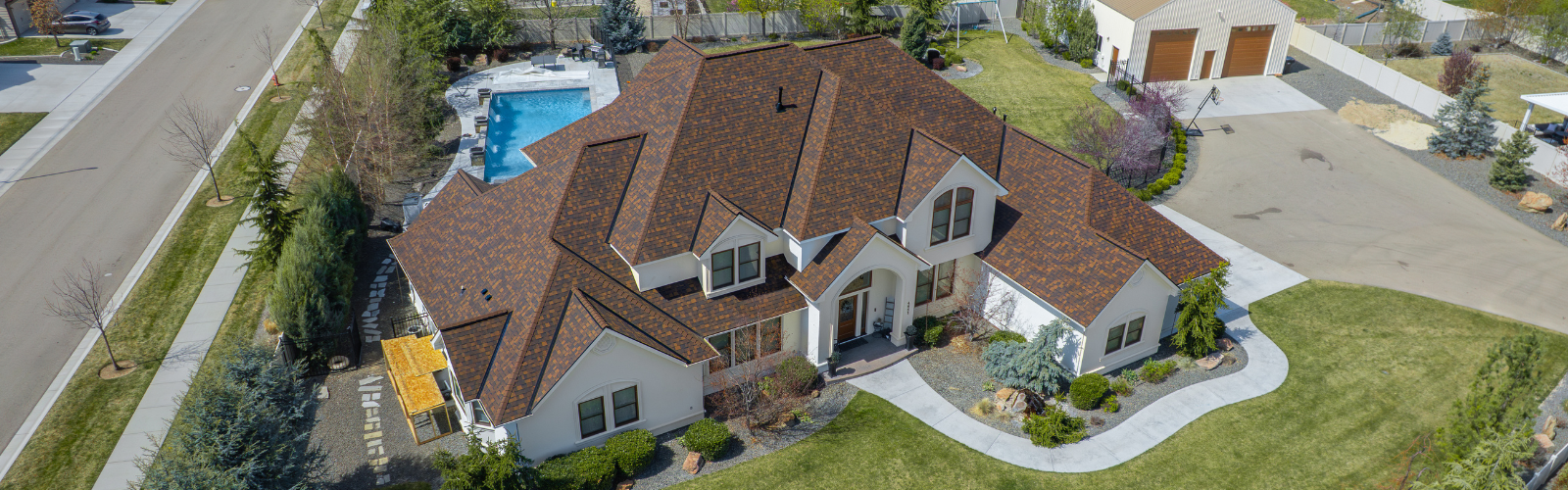 Aerial view of a large home with a clean, well-maintained shingle roof surrounded by manicured landscaping and a backyard pool.