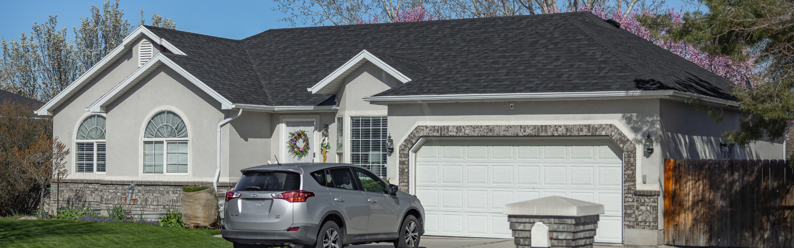 Single-story residential home with a dark asphalt shingle roof, light stucco exterior, and decorative brick accents around the garage. A silver SUV is parked in the driveway, and springtime trees with blooming flowers are visible in the background.