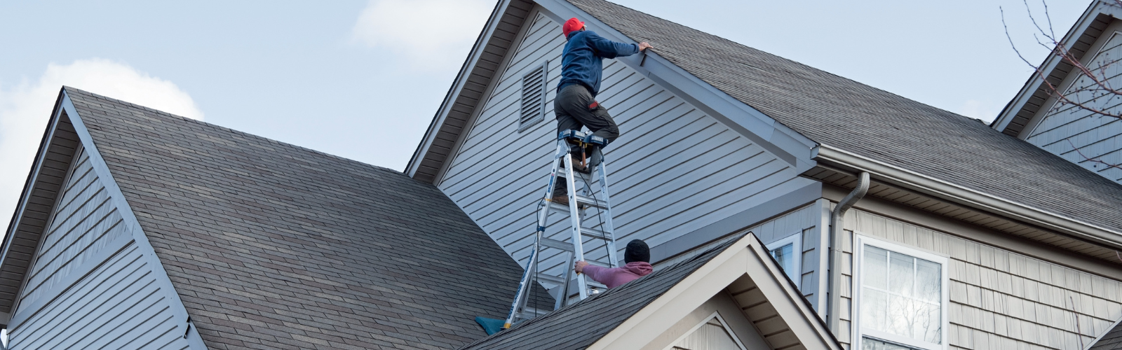 Two workers inspecting a residential roof; one is standing on a ladder examining the roofline while the other stabilizes the ladder from below. The home has asphalt shingles and light gray siding.