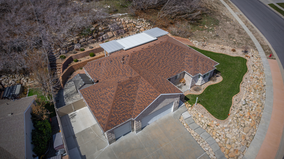 Overhead shot of a suburban home with a new reddish-brown asphalt shingle roof, a neatly manicured lawn, and a curved driveway nestled against a landscaped hillside.