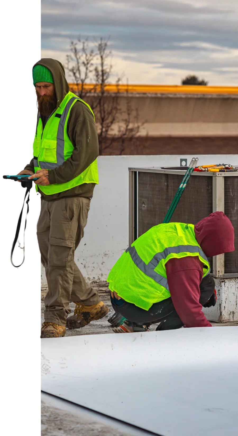 Two commercial roofers in high-visibility vests work on a flat commercial roof. One is standing with a tool in hand while the other kneels near an HVAC unit, surrounded by equipment and supplies, under an overcast sky.