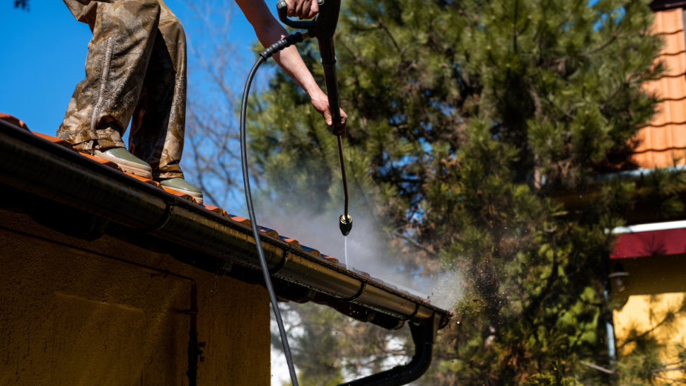 A person standing on a roof uses a pressure washer to clean the gutter, spraying water along the edge while wearing work boots and pants splattered with dirt.