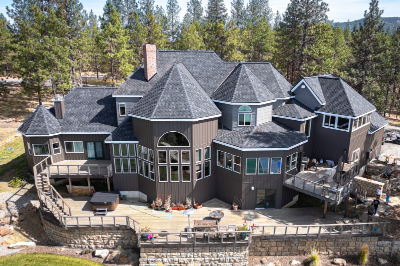 A large, multi-level home with dark gray asphalt shingles, multiple gables, and a spacious wraparound deck, surrounded by tall pine trees.