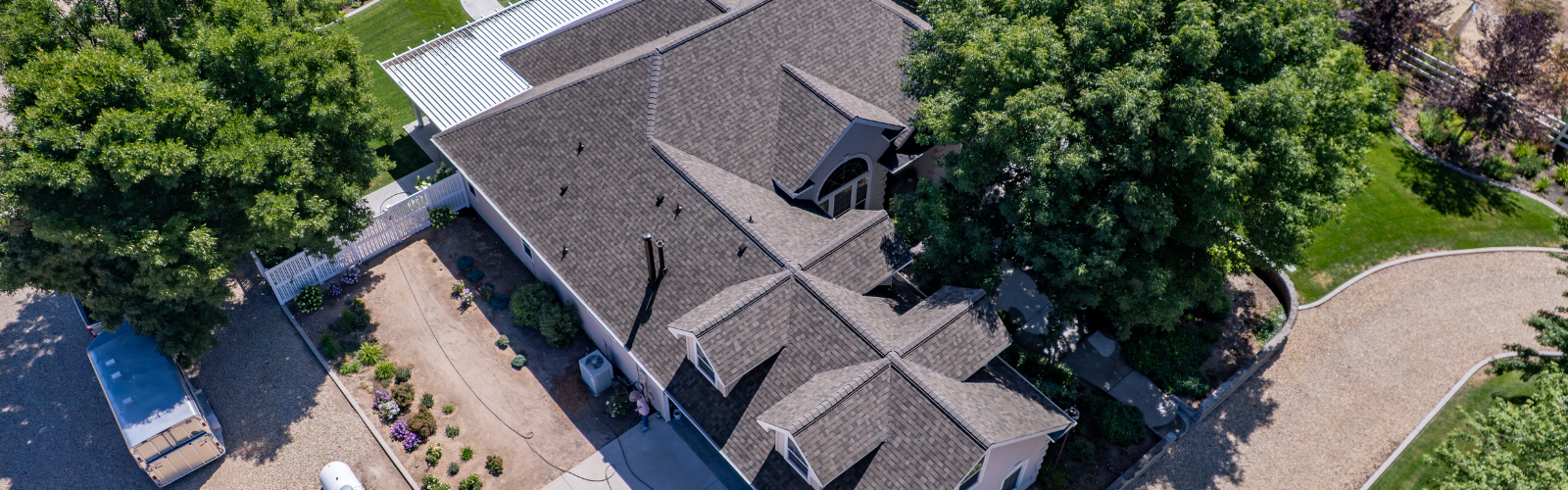 Aerial view of a residential home with a newly installed dark asphalt shingle roof, surrounded by mature trees, landscaped garden beds, and a gravel driveway.