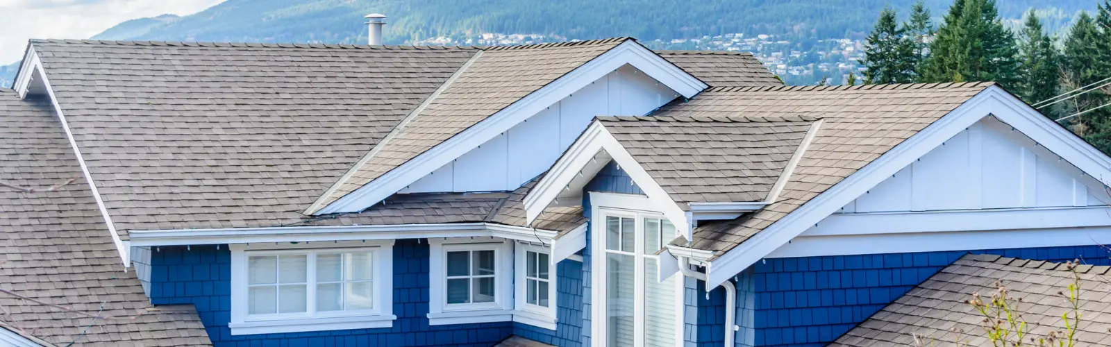 Modern residential roof with gray asphalt shingles on a blue house, surrounded by trees and mountains in the background.