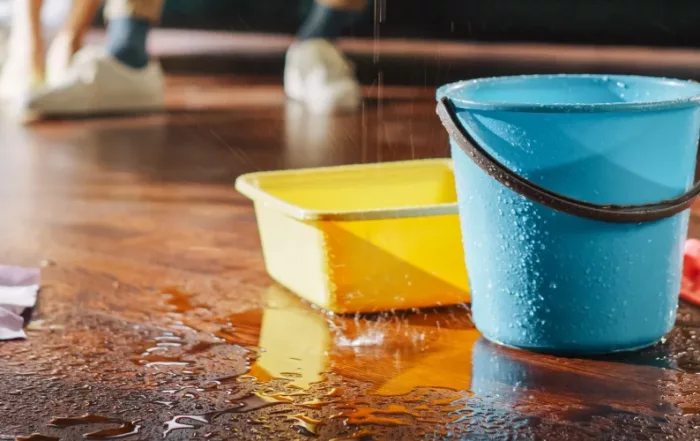 Buckets and rags placed on a wooden floor catching water from a roof leak, with water droplets visible around them.